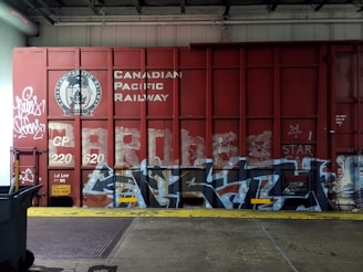 A red freight train car covered with graffiti and the words 'Canadian Pacific Railway' prominently displayed. There are various tags and artistic designs in shades of white and blue on the train car. The setting appears to be inside a warehouse or an industrial area, with a metal roller door and a trash bin visible to the side. The floor consists of concrete with a yellow safety line along the edge.