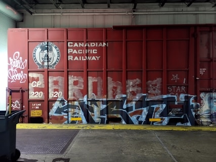 A red freight train car covered with graffiti and the words 'Canadian Pacific Railway' prominently displayed. There are various tags and artistic designs in shades of white and blue on the train car. The setting appears to be inside a warehouse or an industrial area, with a metal roller door and a trash bin visible to the side. The floor consists of concrete with a yellow safety line along the edge.