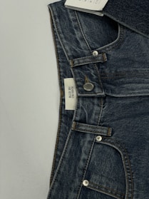 A pair of dark blue denim jeans featuring a visible button and belt loops. There is a tag on the inside waistband with text. The material has a textured appearance characteristic of denim fabric.
