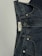 A pair of dark blue denim jeans featuring a visible button and belt loops. There is a tag on the inside waistband with text. The material has a textured appearance characteristic of denim fabric.