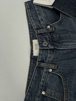 A pair of dark blue denim jeans featuring a visible button and belt loops. There is a tag on the inside waistband with text. The material has a textured appearance characteristic of denim fabric.