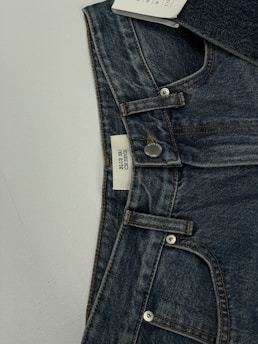 A pair of dark blue denim jeans featuring a visible button and belt loops. There is a tag on the inside waistband with text. The material has a textured appearance characteristic of denim fabric.