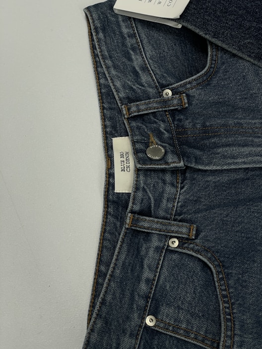 A pair of dark blue denim jeans featuring a visible button and belt loops. There is a tag on the inside waistband with text. The material has a textured appearance characteristic of denim fabric.