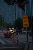 A dimly lit street scene features a small red food truck parked on the side, with its lights on. A solitary person is sitting nearby, eating. Tall buildings with many lit windows form the background, and a prominent yellow warning sign is visible in the foreground, partially obscured by trees.