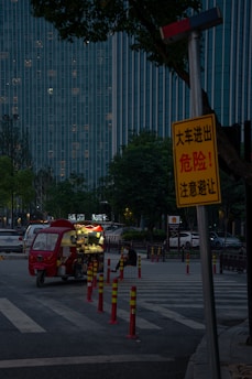 A dimly lit street scene features a small red food truck parked on the side, with its lights on. A solitary person is sitting nearby, eating. Tall buildings with many lit windows form the background, and a prominent yellow warning sign is visible in the foreground, partially obscured by trees.