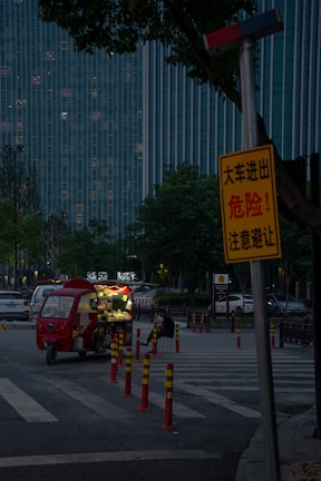 A dimly lit street scene features a small red food truck parked on the side, with its lights on. A solitary person is sitting nearby, eating. Tall buildings with many lit windows form the background, and a prominent yellow warning sign is visible in the foreground, partially obscured by trees.
