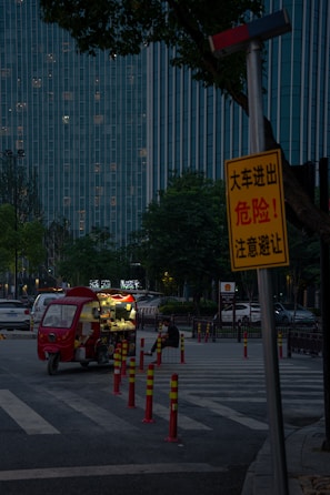 A dimly lit street scene features a small red food truck parked on the side, with its lights on. A solitary person is sitting nearby, eating. Tall buildings with many lit windows form the background, and a prominent yellow warning sign is visible in the foreground, partially obscured by trees.