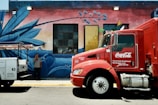 A large red Coca-Cola delivery truck is parked next to a vibrant, painted mural featuring blue agave plants on a brick wall. A man stands on the sidewalk, holding a phone to his ear, partially obscured by a white utility vehicle. Colorful flags hang above the window, enhancing the lively scene.