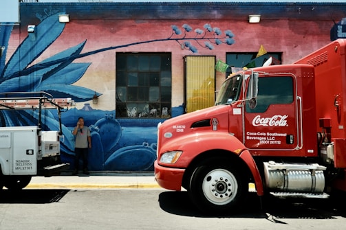 A large red Coca-Cola delivery truck is parked next to a vibrant, painted mural featuring blue agave plants on a brick wall. A man stands on the sidewalk, holding a phone to his ear, partially obscured by a white utility vehicle. Colorful flags hang above the window, enhancing the lively scene.