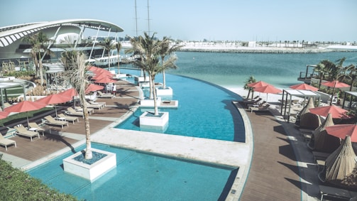 A luxurious outdoor pool area overlooks a calm body of water. Sun loungers with red umbrellas are arranged on a wooden deck, and several palm trees are planted in square planters. A modern architectural building curves gracefully in the background, and the scene is bright and tranquil.
