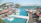 A luxurious outdoor pool area overlooks a calm body of water. Sun loungers with red umbrellas are arranged on a wooden deck, and several palm trees are planted in square planters. A modern architectural building curves gracefully in the background, and the scene is bright and tranquil.