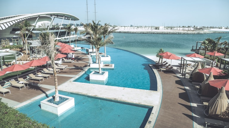 A luxurious outdoor pool area overlooks a calm body of water. Sun loungers with red umbrellas are arranged on a wooden deck, and several palm trees are planted in square planters. A modern architectural building curves gracefully in the background, and the scene is bright and tranquil.