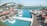 A luxurious outdoor pool area overlooks a calm body of water. Sun loungers with red umbrellas are arranged on a wooden deck, and several palm trees are planted in square planters. A modern architectural building curves gracefully in the background, and the scene is bright and tranquil.