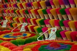 Bundles of incense sticks are arranged in a visually striking pattern, displaying a variety of bright colors including red, yellow, green, purple, and pink. The incense sticks are tied together and fanned out, creating a textured surface with multiple layers. White wicker baskets are placed among the incense sticks, adding to the composition's balance and depth.