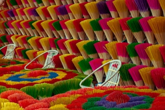 Bundles of incense sticks are arranged in a visually striking pattern, displaying a variety of bright colors including red, yellow, green, purple, and pink. The incense sticks are tied together and fanned out, creating a textured surface with multiple layers. White wicker baskets are placed among the incense sticks, adding to the composition's balance and depth.