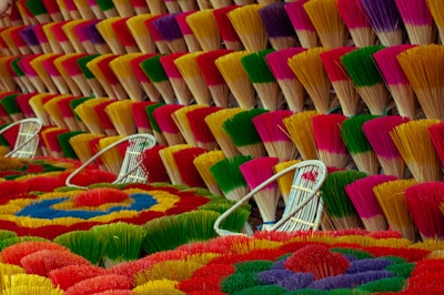 Bundles of incense sticks are arranged in a visually striking pattern, displaying a variety of bright colors including red, yellow, green, purple, and pink. The incense sticks are tied together and fanned out, creating a textured surface with multiple layers. White wicker baskets are placed among the incense sticks, adding to the composition's balance and depth.