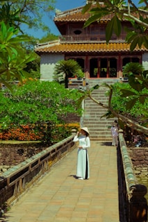 A woman in traditional attire stands on a stone pathway leading to a historical building. The backdrop consists of lush greenery and a vibrant flower garden. The architecture features an ornate roof and multiple columns, reflecting traditional design elements.