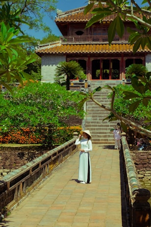 A woman in traditional attire stands on a stone pathway leading to a historical building. The backdrop consists of lush greenery and a vibrant flower garden. The architecture features an ornate roof and multiple columns, reflecting traditional design elements.