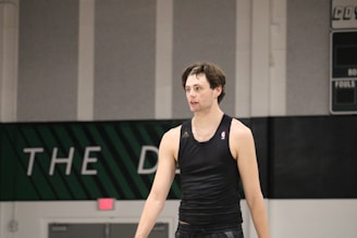 A person wearing a sleeveless black sports top is standing indoors in a gymnasium. The background includes a gray wall with green and white lettering partially visible.