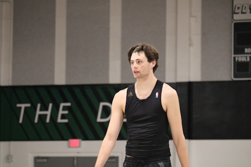 A person wearing a sleeveless black sports top is standing indoors in a gymnasium. The background includes a gray wall with green and white lettering partially visible.