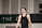 A person wearing a sleeveless black sports top is standing indoors in a gymnasium. The background includes a gray wall with green and white lettering partially visible.