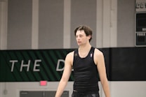 A person wearing a sleeveless black sports top is standing indoors in a gymnasium. The background includes a gray wall with green and white lettering partially visible.