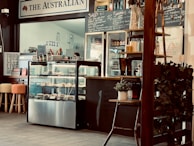 A cozy café interior featuring a counter with a display case filled with pastries. Chalkboards behind the counter list juices, smoothies, and milkshake options. Wooden stools with colorful cushioned seats are arranged in front of the counter. Potted plants and other decorative elements add to the ambiance.