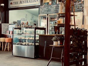 A cozy café interior featuring a counter with a display case filled with pastries. Chalkboards behind the counter list juices, smoothies, and milkshake options. Wooden stools with colorful cushioned seats are arranged in front of the counter. Potted plants and other decorative elements add to the ambiance.