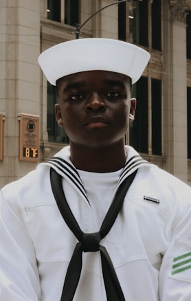 A person wearing a white naval uniform stands in an urban setting with a background of tall buildings and a sign displaying the temperature as 87°F. The uniform includes a traditional sailor hat and a black neckerchief.