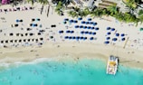 An aerial view of a beach with rows of colorful umbrellas and sunbeds. Palm trees line the back of the beach, and a catamaran is docked on the shore. The turquoise water has people swimming and playing near the shoreline.