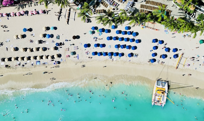 An aerial view of a beach with rows of colorful umbrellas and sunbeds. Palm trees line the back of the beach, and a catamaran is docked on the shore. The turquoise water has people swimming and playing near the shoreline.