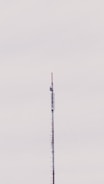 A tall, slender telecommunications tower extends vertically against a pale, overcast sky. The structure, composed of metal and various antenna components, appears to have several platforms and wirings attached to it.