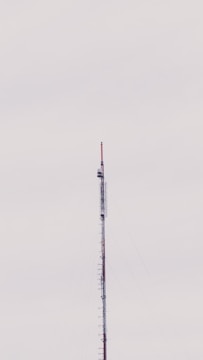 A tall, slender telecommunications tower extends vertically against a pale, overcast sky. The structure, composed of metal and various antenna components, appears to have several platforms and wirings attached to it.