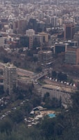 A dense urban skyline filled with a variety of buildings, including high-rise apartments and office buildings, is visible. In the foreground, a large apartment or office building stands amidst lush green trees. A swimming pool can be seen surrounded by trees and lounge chairs near the lower part of the area. Several roads with cars are noticeable, along with construction cranes indicating ongoing development.