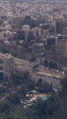 A dense urban skyline filled with a variety of buildings, including high-rise apartments and office buildings, is visible. In the foreground, a large apartment or office building stands amidst lush green trees. A swimming pool can be seen surrounded by trees and lounge chairs near the lower part of the area. Several roads with cars are noticeable, along with construction cranes indicating ongoing development.