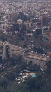 A dense urban skyline filled with a variety of buildings, including high-rise apartments and office buildings, is visible. In the foreground, a large apartment or office building stands amidst lush green trees. A swimming pool can be seen surrounded by trees and lounge chairs near the lower part of the area. Several roads with cars are noticeable, along with construction cranes indicating ongoing development.