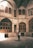 An individual stands in the center of a historical courtyard, capturing a photograph with a smartphone. The architecture features arched windows and intricate details, and the interior is illuminated by soft natural light filtering in from above.