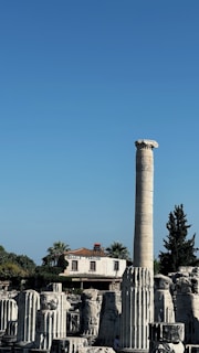 Ruins of ancient columns with a tall, intact column standing prominently in the foreground. A building labeled 'Oracle Pansion' with a terra-cotta roof is visible in the background, surrounded by palm trees. The sky above is clear and blue.