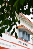 A vintage-style building with the words 'Post Office' on its facade is partially obscured by lush green leaves. The structure features blue shutters and an orange trim against a white exterior, evoking a classic architectural vibe.