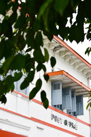 A vintage-style building with the words 'Post Office' on its facade is partially obscured by lush green leaves. The structure features blue shutters and an orange trim against a white exterior, evoking a classic architectural vibe.