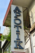 A vertical sign for a pharmacy with the word 'APOTIK' in blue neon letters mounted on the side of a building. The building has a weathered appearance with plants growing nearby, adding a touch of greenery.