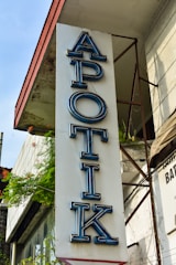 A vertical sign for a pharmacy with the word 'APOTIK' in blue neon letters mounted on the side of a building. The building has a weathered appearance with plants growing nearby, adding a touch of greenery.