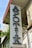 A vertical sign for a pharmacy with the word 'APOTIK' in blue neon letters mounted on the side of a building. The building has a weathered appearance with plants growing nearby, adding a touch of greenery.