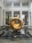 A large, abstract golden sculpture with a hollow center is prominently displayed. It is situated on a polished black platform surrounded by a shallow water feature. The background features a modern architectural setting with high ceilings, columns, and large windows. The area is illuminated by natural light, and there are potted plants in the vicinity.