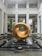 A large, abstract golden sculpture with a hollow center is prominently displayed. It is situated on a polished black platform surrounded by a shallow water feature. The background features a modern architectural setting with high ceilings, columns, and large windows. The area is illuminated by natural light, and there are potted plants in the vicinity.