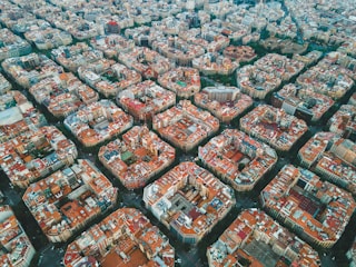 A densely populated urban area seen from above, characterized by a grid-like pattern of blocks with buildings arranged in a consistent and organized fashion. The buildings predominantly have reddish-brown rooftops, with a mix of higher and lower structures. Streets are visible in between the blocks, forming somewhat square-shaped sections. Greenery and parks can be seen in some parts of the image, providing contrast to the dense urban environment.