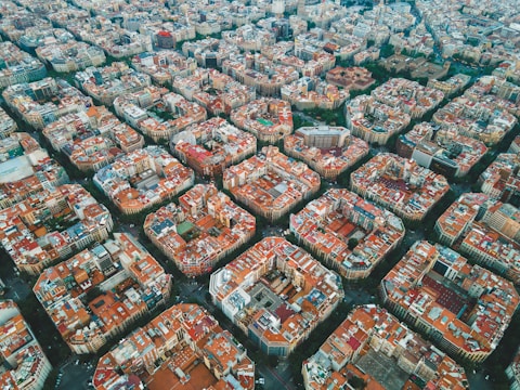 A densely populated urban area seen from above, characterized by a grid-like pattern of blocks with buildings arranged in a consistent and organized fashion. The buildings predominantly have reddish-brown rooftops, with a mix of higher and lower structures. Streets are visible in between the blocks, forming somewhat square-shaped sections. Greenery and parks can be seen in some parts of the image, providing contrast to the dense urban environment.
