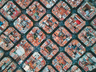 Aerial view of an urban area with a grid-like pattern of residential buildings, each block forming an octagonal shape. The rooftops are predominantly red with some scattered beige and white structures. Streets intersect neatly at corners, showcasing a well-planned infrastructure.