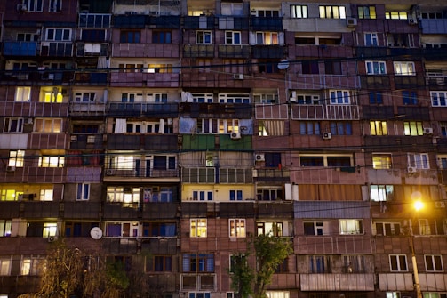 A multi-story apartment building with numerous balconies and windows, many of which are illuminated. The building displays a mix of colors and materials, with balconies enclosed in glass or metal and various curtains visible. Some trees and streetlights are visible at the bottom.