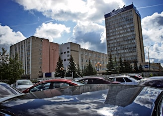 A large multi-story building with numerous windows stands against a partly cloudy sky. In front of the building, there are several evergreen trees and a parked car lot filled with vehicles. There is a reflection of the cloudy sky on the roof of one of the cars.